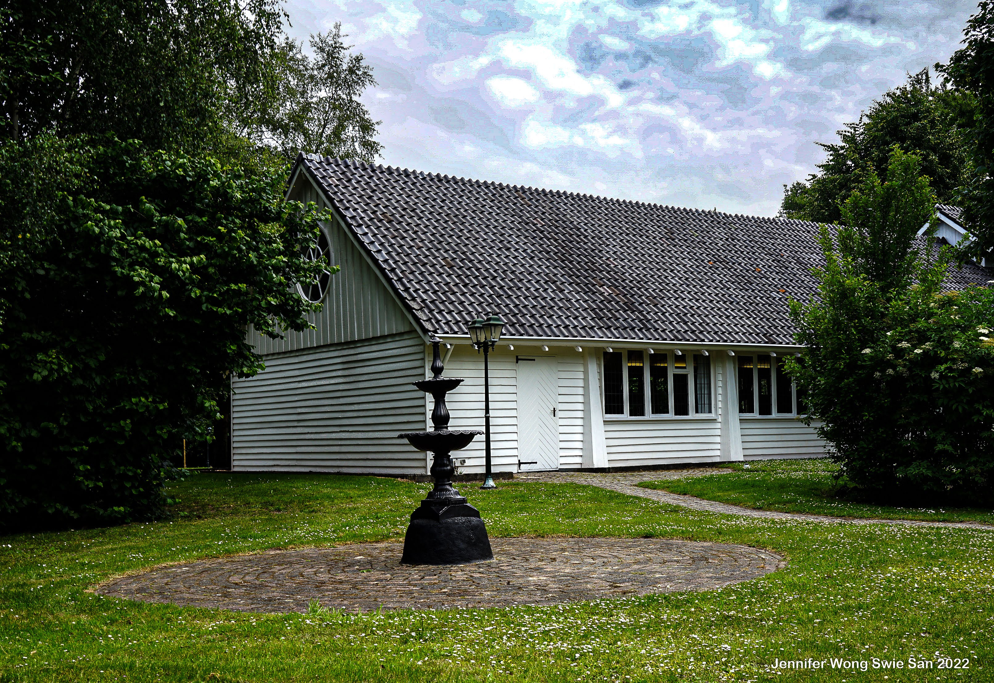Wit kerkje met gietijzeren fontein op de voorgrond en dramatische wolkenlucht